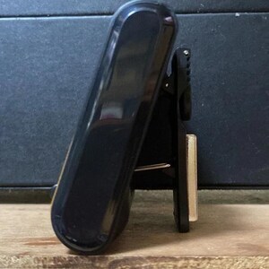 Pode incluir: Uma presilha preta e brilhante com uma parte superior arredondada e um design articulado. A presilha est&aacute; posicionada em &acirc;ngulo, revelando seu perfil lateral e um pequeno componente met&aacute;lico retangular. O fundo &eacute; uma superf&iacute;cie escura e texturizada.