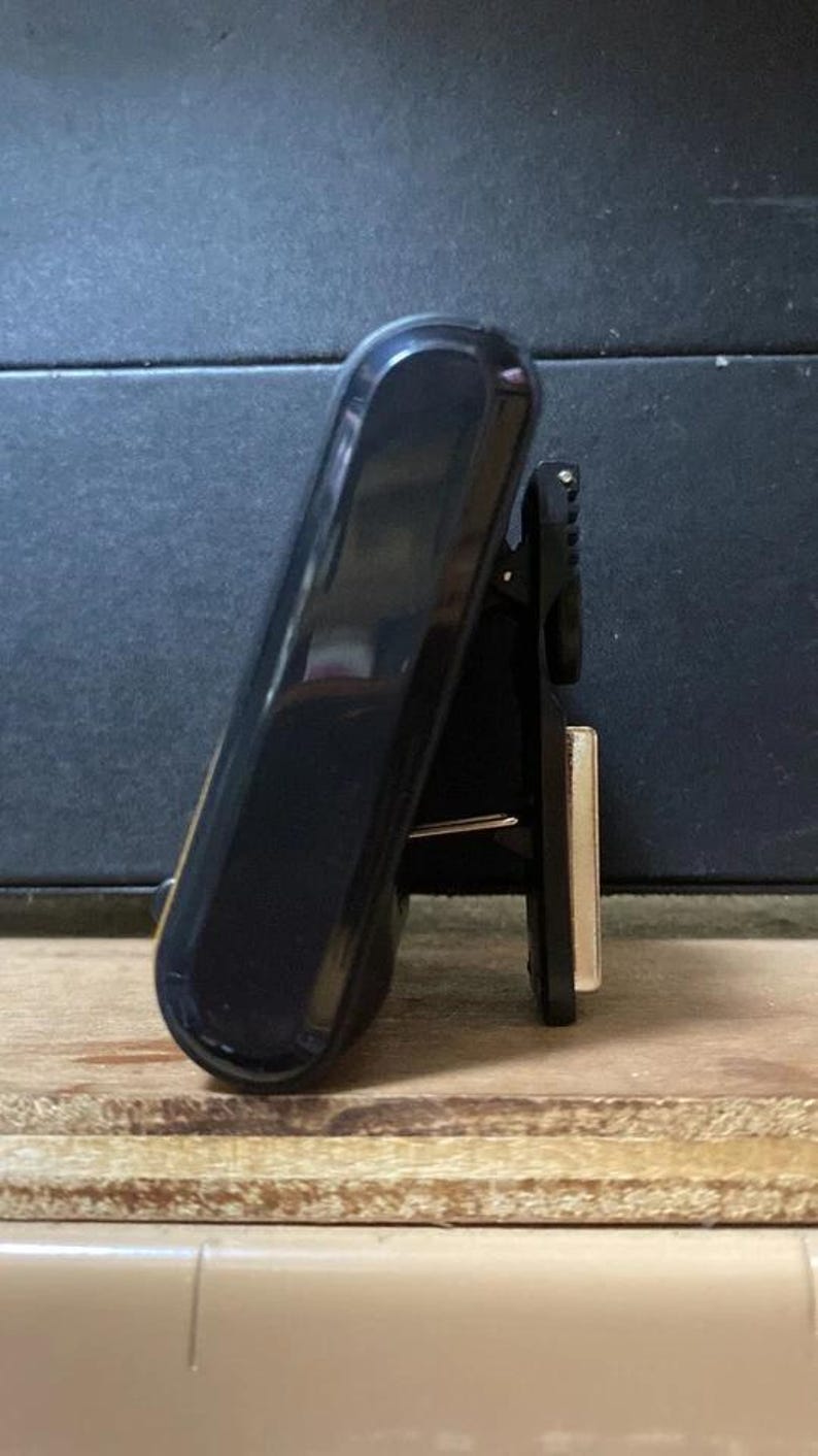 Pode incluir: Uma presilha preta e brilhante com uma parte superior arredondada e um design articulado. A presilha est&aacute; posicionada em &acirc;ngulo, revelando seu perfil lateral e um pequeno componente met&aacute;lico retangular. O fundo &eacute; uma superf&iacute;cie escura e texturizada.