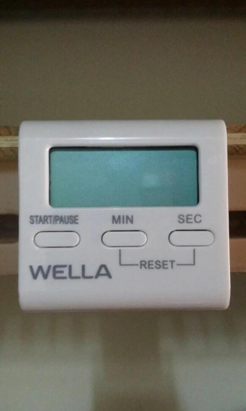 Pode incluir: Um temporizador de cozinha digital branco com um visor retangular e tr&ecirc;s bot&otilde;es rotulados como "START/PAUSE", "MIN" e "SEC". A marca "WELLA" est&aacute; impressa na frente. O temporizador tem um bot&atilde;o "RESET".