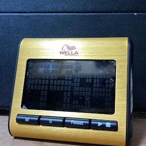 May include: A gold-colored digital timer with a black display screen. The timer has a rectangular shape with rounded corners and black buttons labeled "M", "S", and "Reset". The brand "WELLA PROFESSIONALS" is printed at the top.