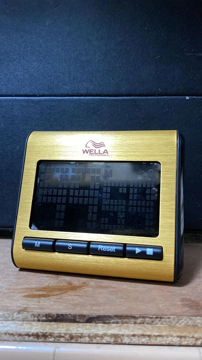 Pode incluir: Um temporizador digital dourado com tela preta. O temporizador tem formato retangular com cantos arredondados e bot&otilde;es pretos com as etiquetas "M", "S" e "Reset". A marca "WELLA PROFESSIONALS" est&aacute; impressa no topo.