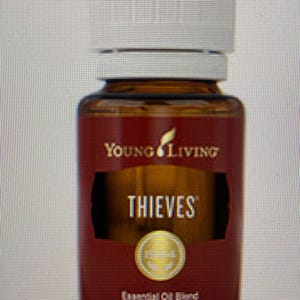 Op de afbeelding: Een bruine glazen fles met een witte dop, met het opschrift "Young Living Thieves Essential Oil Blend, 15 ml." Het etiket is bordeauxrood met gouden letters en een gouden embleem. De fles bevat een goudkleurige vloeistof.