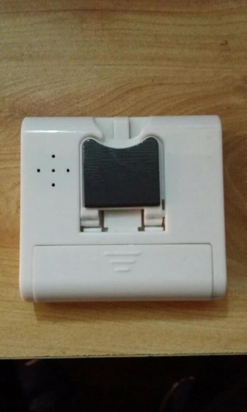 Pode incluir: Um dispositivo eletr&ocirc;nico retangular branco com uma se&ccedil;&atilde;o retangular preta no centro. O dispositivo possui uma s&eacute;rie de pequenos orif&iacute;cios no lado esquerdo e um mecanismo deslizante na parte inferior. &Eacute; provavelmente um pequeno gadget eletr&ocirc;nico.