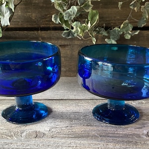 May include: Two cobalt blue glass dessert bowls with a rounded bowl shape and a short stem. The bowls are made of a transparent glass, with a gradient of color from a deep blue to a lighter blue. The bowls are sitting on a wooden surface.