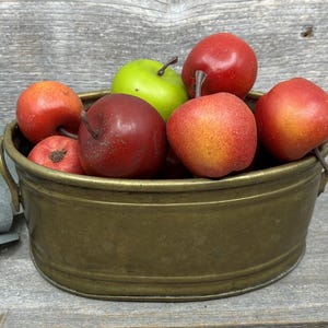 May include: An oval brass basin with two side handles filled with a collection of artificial fruit, including several red apples and one bright green apple, each featuring a small brown stem attached to the top of the fruit.