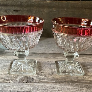 May include: Two clear glass dessert cups with a ruby red rim. The cups have a diamond-cut pattern and sit on a square base. The background is a weathered wooden surface.