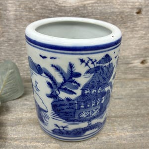 May include: A cylindrical ceramic vessel featuring a white base with intricate blue hand-painted illustrations of traditional pagodas, lush trees, and mountainous landscapes. The rim and base are accented with solid blue horizontal bands.