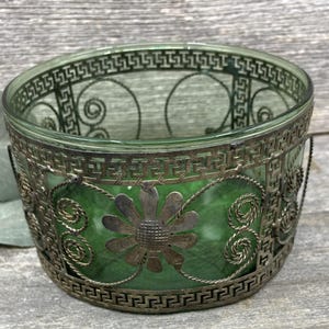 May include: A decorative glass votive candle holder with a green interior and a silver-colored metal filigree overlay. The metalwork features geometric and floral designs. The candle holder is approximately 3.5 inches in diameter.