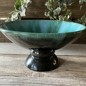 May include: A pedestal bowl with a wide, shallow basin. The bowl's interior transitions from teal to dark green, with a glossy finish. The base is a solid, dark green color. The bowl is likely made of ceramic or glass.