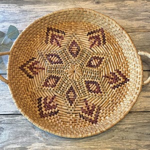 May include: A round woven basket tray with two side handles, featuring a natural tan reed base with a geometric star pattern in shades of dark brown and maroon. The basket has a flat, circular design with a tight, intricate weave throughout.