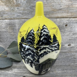 May include: A yellow ceramic jar with a tall neck and a lid. The jar is decorated with a black and white forest scene, including trees and a cabin. The jar is approximately 6 inches tall.