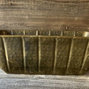 May include: A rectangular, antique brass planter with a hammered texture and vertical ribbed design. The planter has a slightly flared rim and is set against a weathered wooden background.