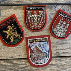 May include: Four embroidered patches with red borders. One patch features a gold lion and the word "HEIDELBERG". Another displays an eagle and the text "FRANKFURT/M". A third patch shows a castle and the word "HAMBURG". The last patch has a building and the text "HOFBRAUHAUS MUNCHEN".