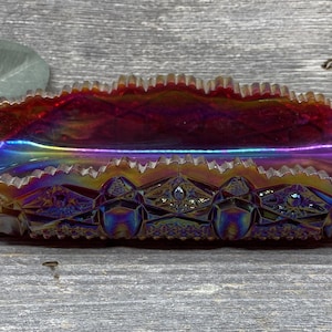 May include: An iridescent oval glass dish with a ruffled rim. The dish has a vibrant color gradient, transitioning from red to purple and gold. The base features a geometric pattern.