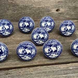 May include: A set of eight round ceramic cabinet knobs featuring a white base with a blue transferware-style pattern depicting a landscape scene with trees and foliage. Each knob has a smooth, glossy finish and a circular shape.