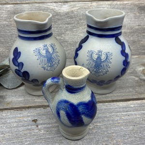 May include: Three gray stoneware pitchers with blue painted details. Two larger pitchers feature a blue eagle emblem and blue leaf patterns around the body. One smaller pitcher has a blue swirl design and a single handle on the side.