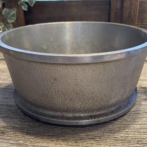 May include: A silver-colored metal bowl with two handles. The bowl has a textured exterior and a wide rim. The bowl sits on a circular base. The background is a wooden surface.
