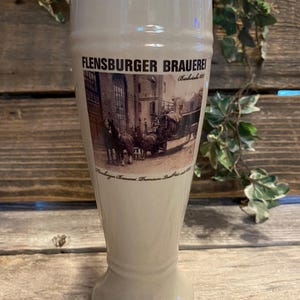 May include: A tall, off-white beer glass with a flared top and a footed base. The glass features the text "FLENSBURGER BRAUEREI" and a sepia-toned image of a horse-drawn carriage. The glass is likely for beer.