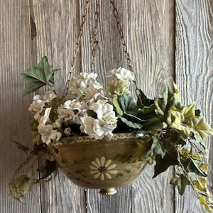 May include: A hanging planter bowl in a muted olive green finish with a painted white daisy motif and scalloped rim. The bowl is suspended by three thin metal chains and is filled with white hydrangea-like flowers and trailing green ivy leaves.