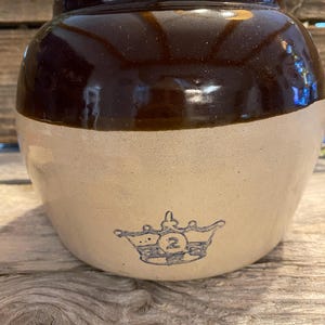 May include: A two-tone stoneware crock with a dark brown glazed top and a tan base. A blue crown with the number 2 is printed on the base. The antique jar is sitting on a wooden surface.