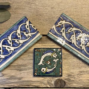 May include: Three ceramic tiles with intricate designs. Two rectangular tiles feature a blue and white pattern with a teal border. A square tile in the center depicts two white fish on a teal background. The tiles are arranged on a wooden surface.