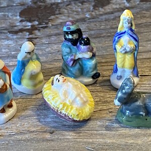 May include: A collection of small, hand-painted ceramic figurines depicting a nativity scene. The figures include Mary, Joseph, baby Jesus in a manger, a lamb, and the three wise men. The figurines are painted in bright colors.