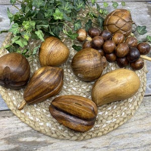 May include: A collection of wooden decorative items, including various shapes like acorns, a pinecone, and a grape cluster, arranged on a woven mat. The items are in shades of brown and are accompanied by green foliage.