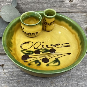 May include: A yellow ceramic olive serving dish with a green rim, featuring the word "Olives" and olive branch illustrations. Includes two small matching containers, one labeled "Nouaux" and the other "Piques".