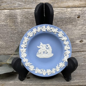 May include: A small, light blue decorative plate with a white relief design. The plate features a central scene of figures and a border of grapevines. The plate is displayed on a black stand against a wooden background.