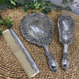 May include: An antique vanity set featuring a hand mirror, a hairbrush, and a comb. The silver-toned pieces have ornate floral and scrollwork designs. The comb has a cream-colored plastic insert.