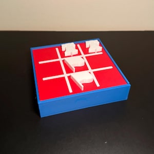 May include: A square tic-tac-toe game board with a blue frame and a red playing surface featuring white grid lines. The board includes four white game pieces shaped like Liberty Bells and flags placed within the grid squares on the red surface.