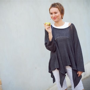 May include: A woman wearing a grey, loose-fitting sweater with a white vest underneath. She is holding a green apple in her hand and smiling.