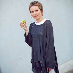 May include: A woman wearing a dark grey, loose-fitting sweater with a white undershirt and white and blue striped shorts. She is holding a green apple in her hand.