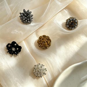 May include: Five decorative buttons featuring intricate beadwork. One is a dark gray starburst, one is a metallic bronze cluster, one is a gold spiral, one is a black flower with clear accents, and one is a white pearl-like sphere with silver details.