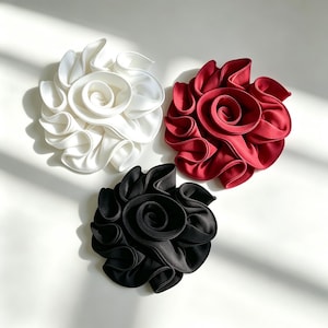 Puede incluir: Tres accesorios para el cabello con forma de flor de tela dispuestos en grupo. Una flor es blanca, otra es de color rojo intenso y otra es negra. Cada pieza presenta pétalos superpuestos y con volantes que se enroscan hacia el centro.