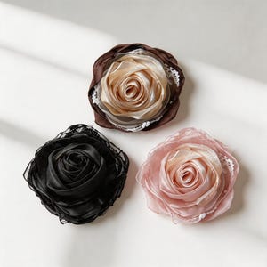 Peut inclure: Trois broches en forme de fleur en tissu avec des pétales en satin superposés et une bordure en dentelle délicate. Une broche est noire, une est beige champagne avec une couche extérieure marron foncé, et une est rose poudré. Forme circulaire.