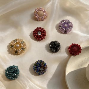 May include: A collection of eight circular, handcrafted beaded buttons in various colors including peach, purple, red, gold, black, dark red, teal, and a multi-colored blue and green mix, each featuring intricate beadwork and central accents.