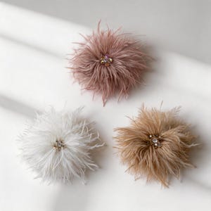 May include: Three circular decorative accessories made of soft, wispy feathers in shades of dusty rose, white, and tan. Each piece features a central cluster of small, sparkling clear and iridescent crystals arranged in a floral-like pattern.