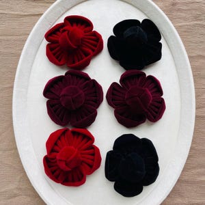 Op de afbeelding: Zes fluwelen bloemvormige ringdozen in tinten rood, bordeauxrood en zwart. De dozen zijn gerangschikt op een wit ovaal dienblad. De bloemblaadjes zijn zichtbaar en het midden van elke bloem is een kleine, ronde vorm.