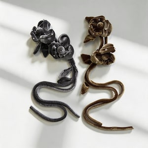 May include: Two long, flexible velvet floral accessories, one in charcoal gray and one in olive brown. Each piece features two sculpted velvet flowers adorned with small, clear crystals, attached to a long, slender, matching velvet-covered stem.