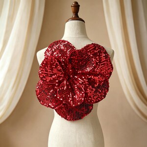 May include: A large, dimensional flower applique crafted from vibrant red sequins. The flower features multiple overlapping, rounded petals that create a full, layered appearance. The entire surface is covered in reflective red sequins.