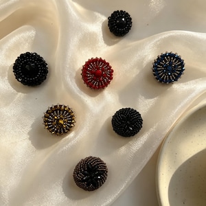 May include: A collection of seven circular buttons featuring intricate beadwork and central faceted stones. The buttons are arranged in various colors including black, red, blue, gold, and bronze, each displaying a detailed radial pattern of small beads.