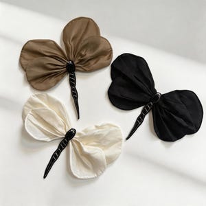 May include: Three dragonfly-shaped hair accessories featuring sheer, pleated fabric wings in tan, black, and off-white. Each piece has a central black body adorned with small, dark beads and a long, slender tail section with white stitched detailing.