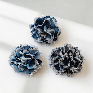 Puede incluir: Tres flores de tela circulares hechas de material de mezclilla azul oscuro. Cada flor presenta múltiples capas de pétalos con bordes deshilachados de color claro, creando una apariencia texturizada y con volantes. La tela tiene un tejido distintivo.