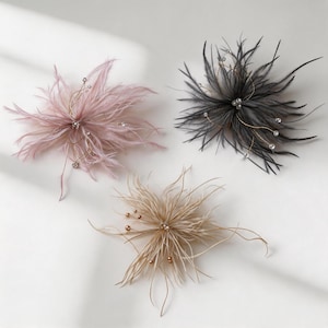 May include: Three decorative floral-shaped hair accessories made of soft feathers. One is light pink, one is black, and one is beige. Each piece features thin gold-toned wire stems extending from the centre, tipped with small white or metallic beads.