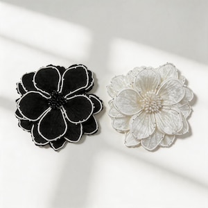 Puede incluir: Dos broches en forma de flor, uno negro y otro blanco. Cada broche presenta pétalos superpuestos con un fino ribete blanco en los bordes y un denso grupo de pequeñas cuentas en el centro. Los pétalos tienen una superficie texturizada y redondeada.