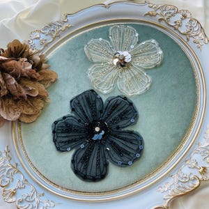 May include: Two decorative flower embellishments. One is a sheer, off-white flower with a silver sequin center. The other is a dark gray flower with black beaded accents and a dark sequin center. Both are on a light green surface.