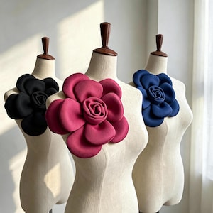 Peut inclure: Trois mannequins en tissu beige présentant chacun une grande fleur décorative en relief. Le mannequin de gauche porte une fleur noire, celui du centre une fleur magenta foncé et celui de droite une fleur bleu foncé.