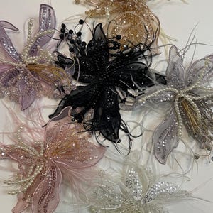 May include: A collection of decorative floral embellishments in various colours. The flowers feature intricate designs with beads, sequins, and feathers. Colours include black, pink, lavender, silver, and gold. These embellishments are ideal for adding a touch of elegance to apparel or accessories.