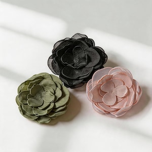 Può includere: Tre accessori a forma di fiore in tessuto disposti in gruppo. Un fiore è nero, uno è verde oliva e uno è rosa pallido. Ogni fiore presenta molteplici petali arrotondati e sovrapposti con bordi cuciti, creando un aspetto voluminoso e pieno.